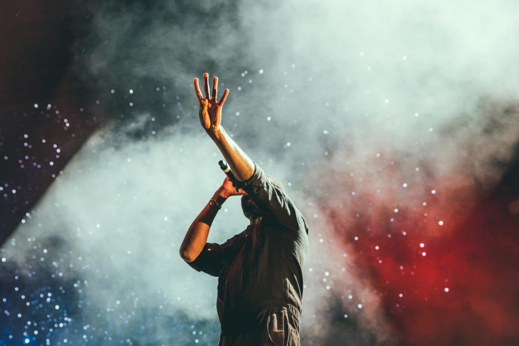 Valokuva laulajasta lavalla, takana savua ja kipinöitä. Kuvan lähde: Austin Neill, Unsplash
