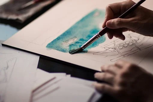Valokuvassa on sivellintä pitelevä käsi, joka maalaa vesivärillä turkoosia merimaisemaa. Maalauksessa lyijykynäluonnoksia veneistä