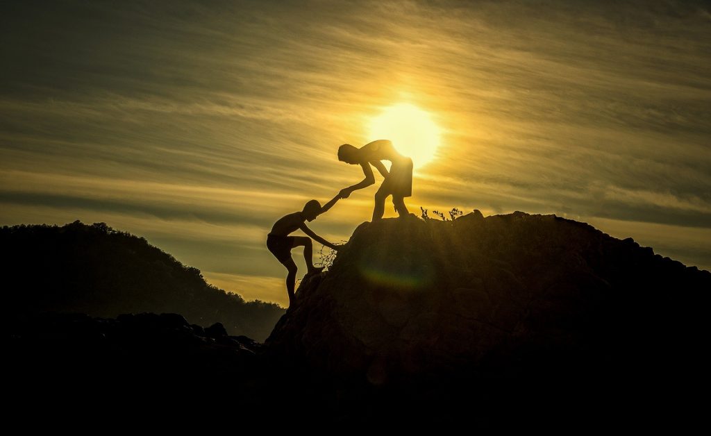 Valokuva, jossa ihminen auttaa toista kiipeämään vuorelle siluettina aurinkoa vasten.
